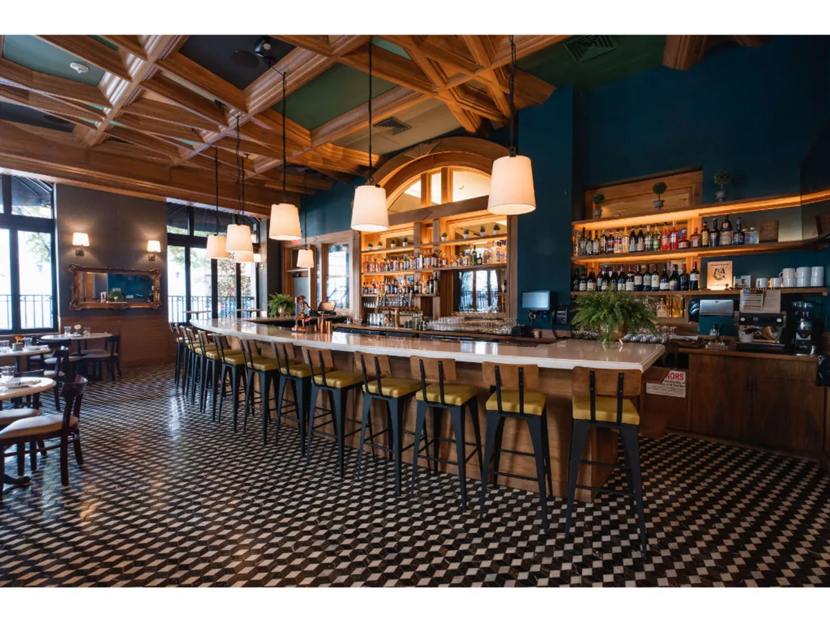 A lively bar scene featuring a long, stylish bar counter topped with bottles and glassware, set beneath an intricately designed ceiling. The bar is surrounded by comfortable seating and unique floor tiles, with subtle lighting and a welcoming atmosphere.
