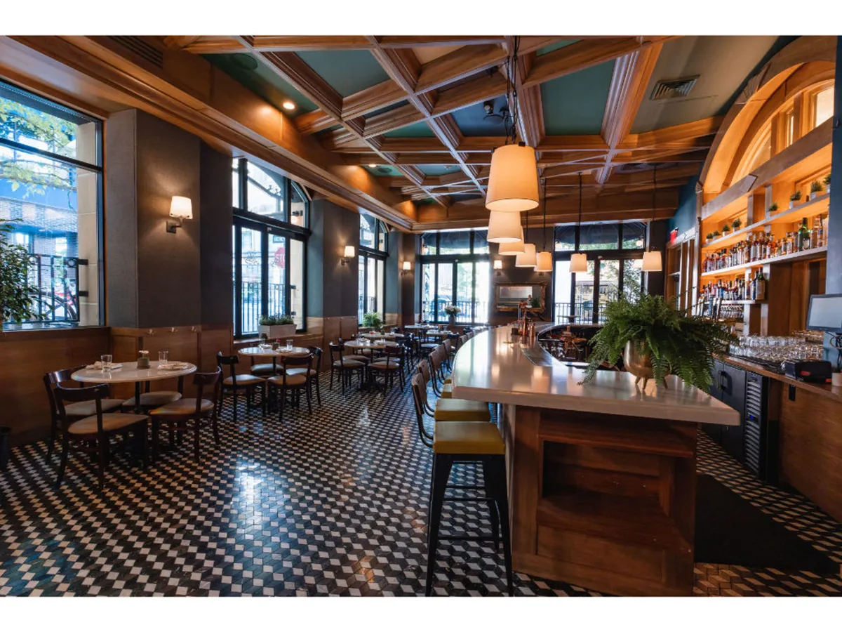 A sophisticated yet comfortable bar and dining space with a stylish bar counter, dining tables, decorative ceiling accents, windows, and lush plants. Guests can be seen socializing, dining, and even working.
