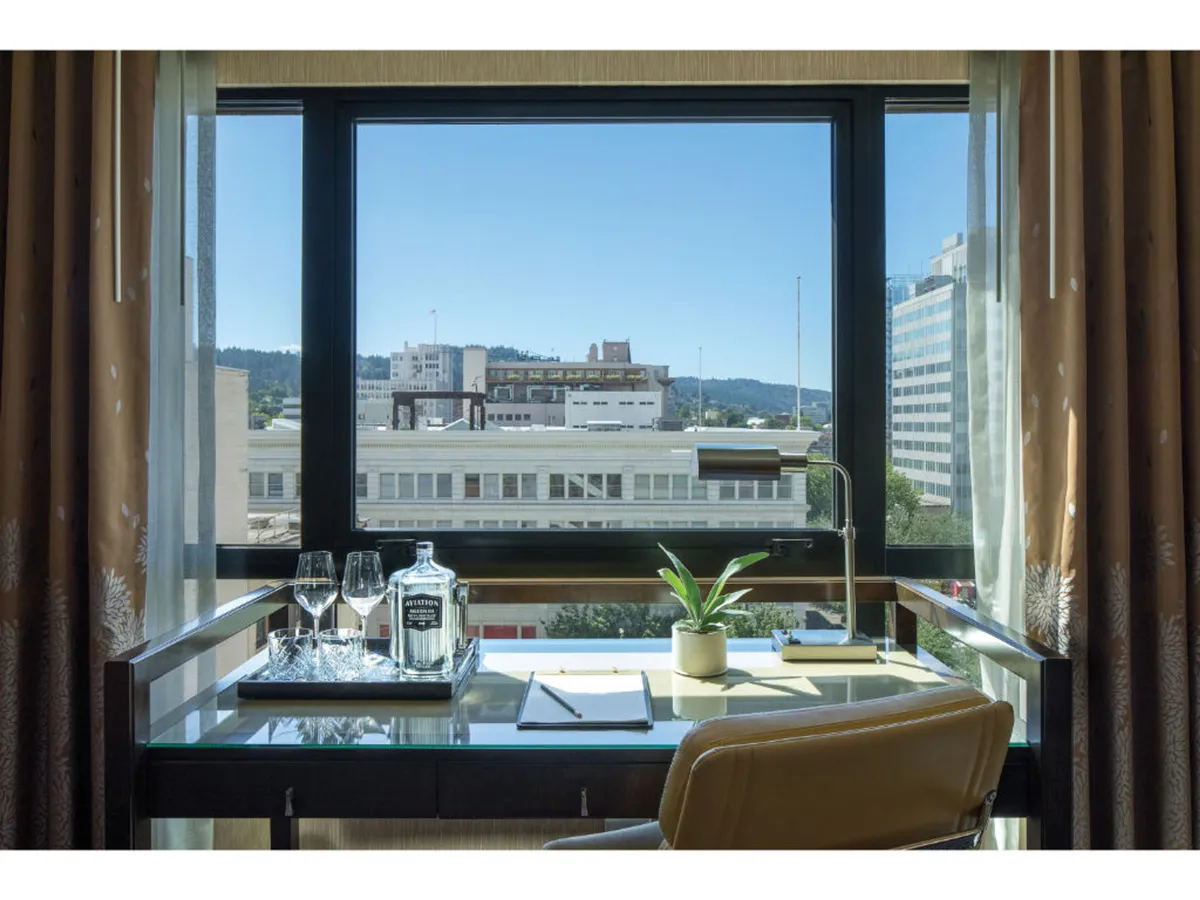 This image features a stylish hotel room with a glass-top desk, a bottle of alcohol with glasses ready for an evening toast, a vibrant green plant adding a touch of nature, and expansive windows revealing stunning city views at sunset. The setup is inviting for work or relaxation, all with Portland's skyline as the backdrop.