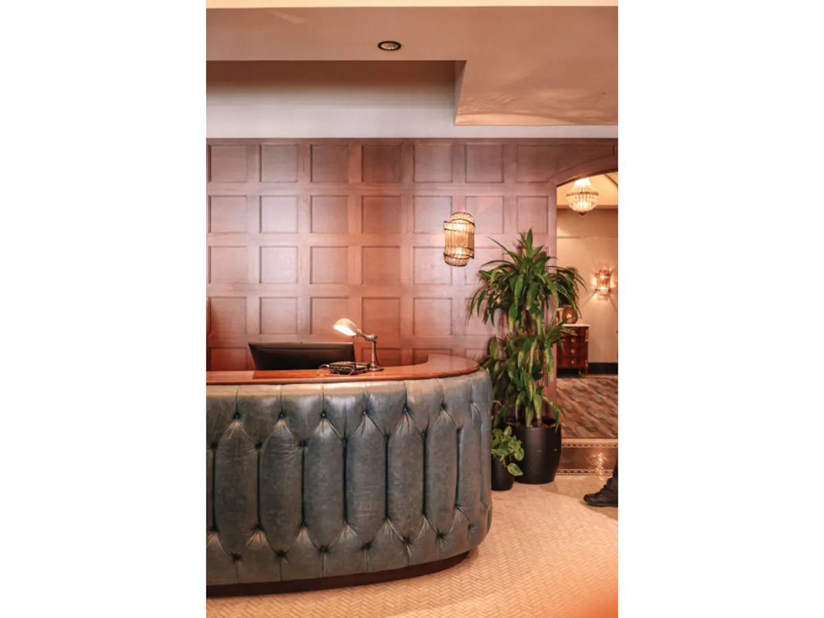 A modern and elegant lobby scene with a curved reception desk, wood-paneled wall, stylish hanging light fixtures, and a potted plant. A staff member is ready behind the desk, creating a welcoming environment for arriving guests.