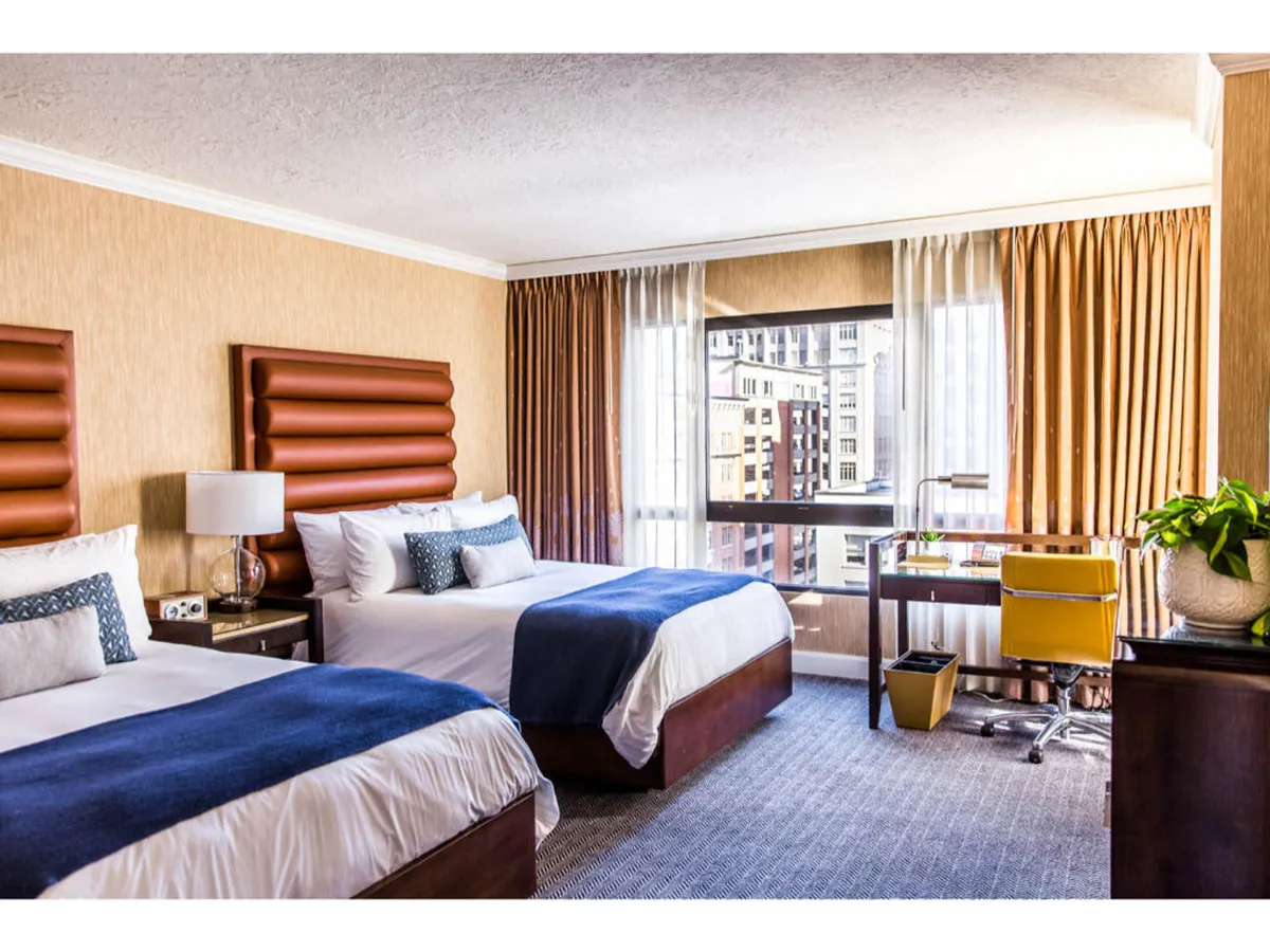 This image features two double beds with stylish blue throws, set against a large window that opens up to sweeping city views. A modern desk with a yellow chair sits by the window, creating a versatile space for work or relaxation. The natural light floods the room, highlighting the comfortable and modern décor.