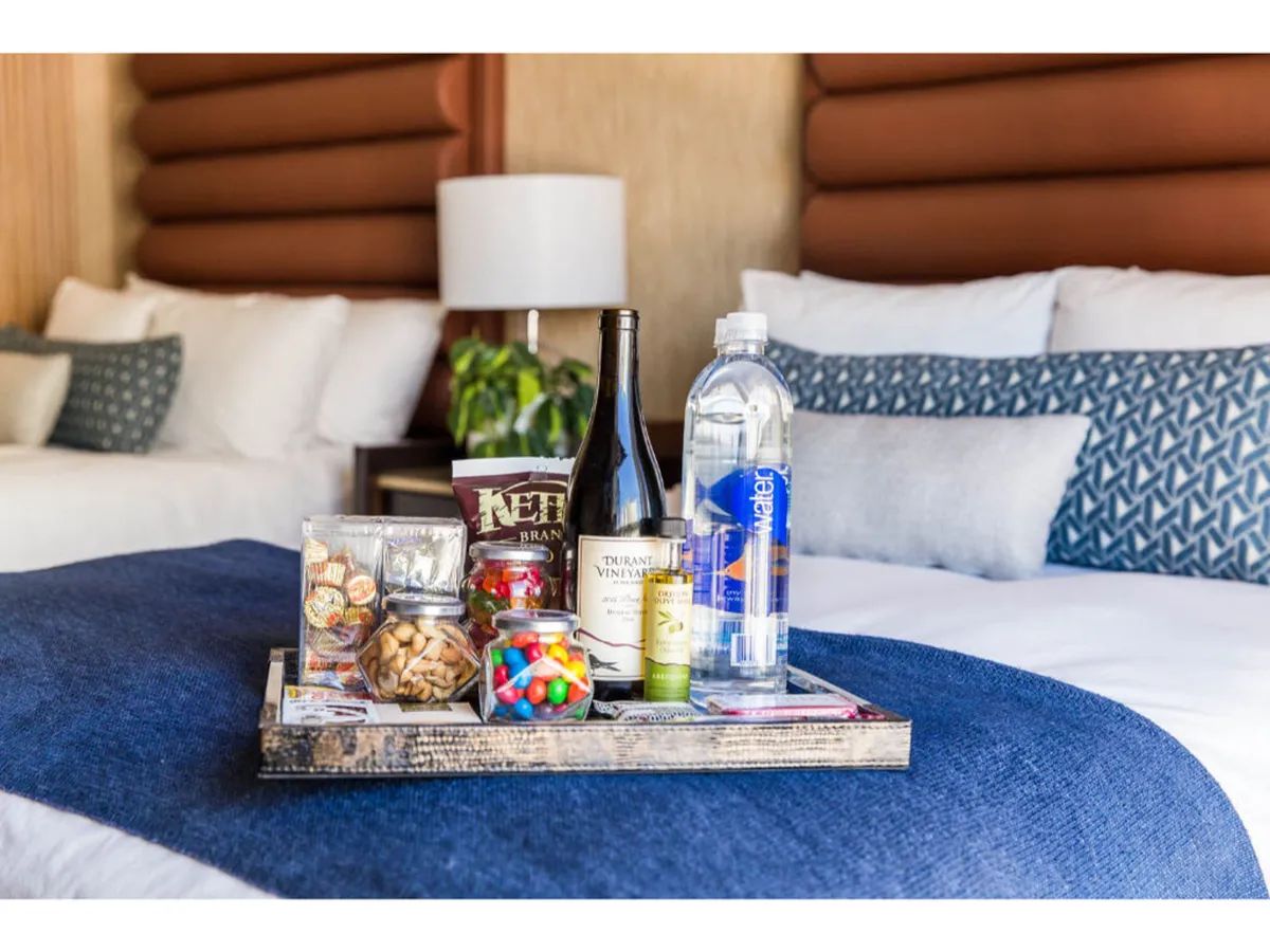 This image shows a cozy bed adorned with decorative pillows, a tray filled with snacks and beverages, and a warm bedside lamp. The inviting setup is perfect for unwinding, enjoying treats, and perhaps watching TV or reading in bed.