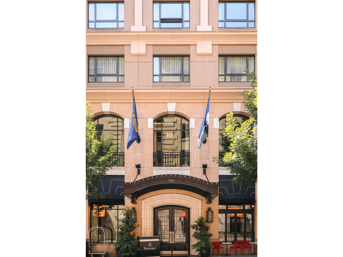 The image showcases the hotel's exterior entrance, featuring elegant stonework, large entrance doors, flags, and lush planters. The scene captures visitors arriving at the property, hinting at both the luxury awaiting inside and the vibrant urban environment just outside.