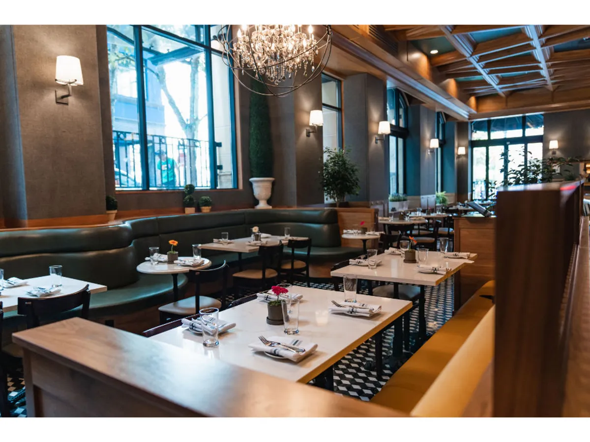 A sophisticated dining space with elegant green seating, a sparkling chandelier overhead, large windows letting in natural light, and expertly set tables, creating an ambiance ideal for socializing, dining, and celebrating.